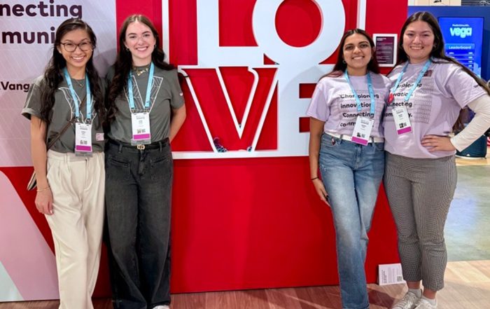 Four women standing in from of a red ‘LOVE’ backdrop at a conference, posing and smiling, wearing badges and lanyards.