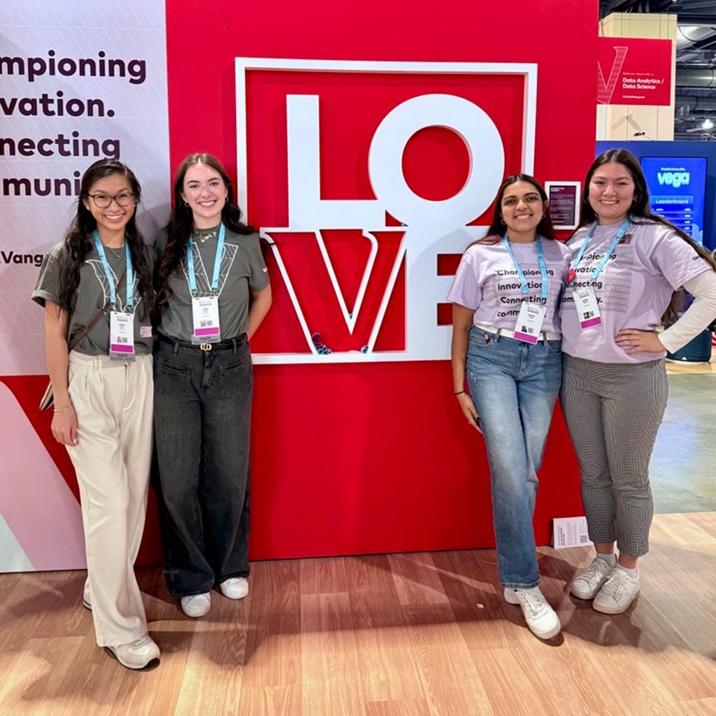 Four women standing in from of a red ‘LOVE’ backdrop at a conference, posing and smiling, wearing badges and lanyards.