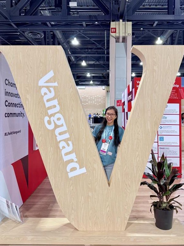 Justina stands behind a large wooden ‘V’ with ‘Vanguard’ written on the side at an indoor conference. She is smiling with her mouth open, with banners and a plant in the background.