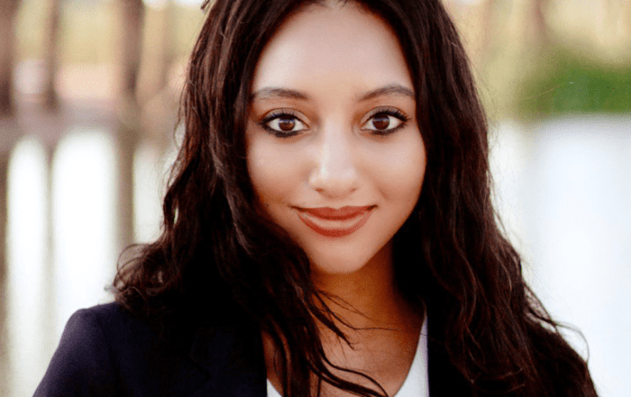 Briana R., wearing a black blazer over a white top, stands outdoors near a calm body of water with trees in the background.