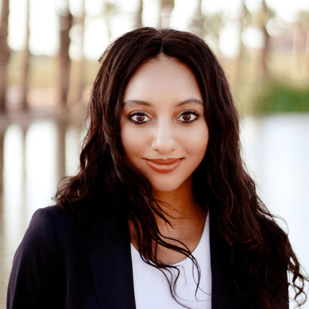 Briana R., wearing a black blazer over a white top, stands outdoors near a calm body of water with trees in the background.