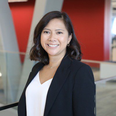 Joy U. smiling in a modern office setting with red and white walls and glass railings, wearing a black blazer over a white top.