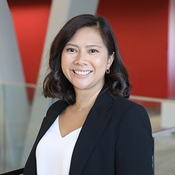 Joy U. smiling in a modern office setting with red and white walls and glass railings, wearing a black blazer over a white top.
