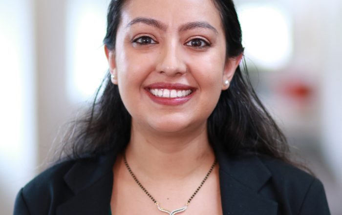 Perryne D. smiling confidently in a green shirt and black blazer. She is against a blurred background of a hallway in an office setting.
