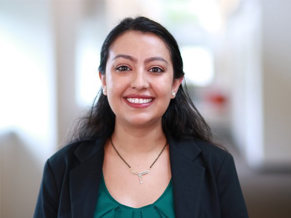 Perryne_Headshot_Edited (1) Joy U. smiling in a modern office setting with red and white walls and glass railings, wearing a black blazer over a white top.