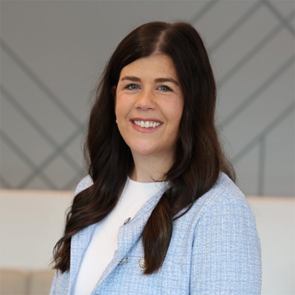Ann D. wearing a light blue textured blazer over a white top, standing indoors against a gray wall with a geometric line pattern.