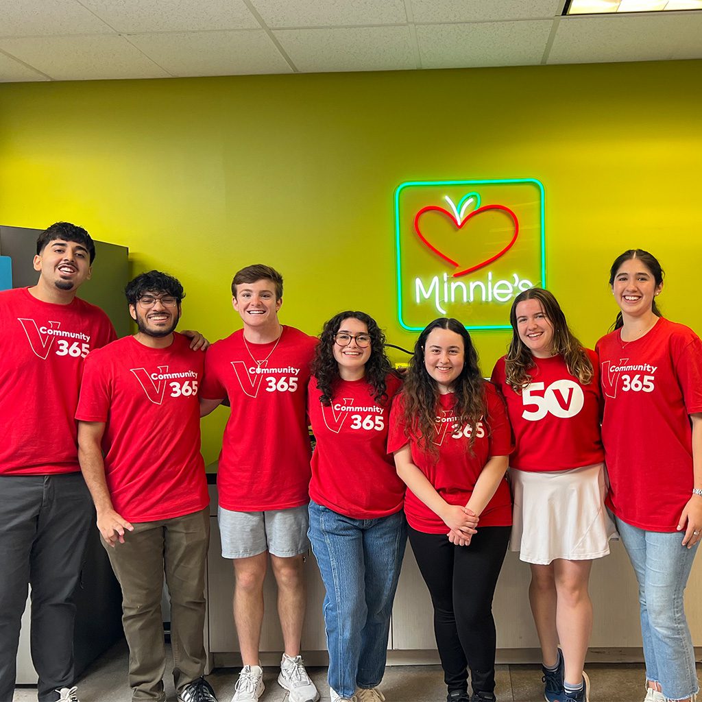 Seven interns with matching red shirts with “Community 365” and “50” printed on them, standing in front of a green wall with a neon sign that says “Minnie’s.”