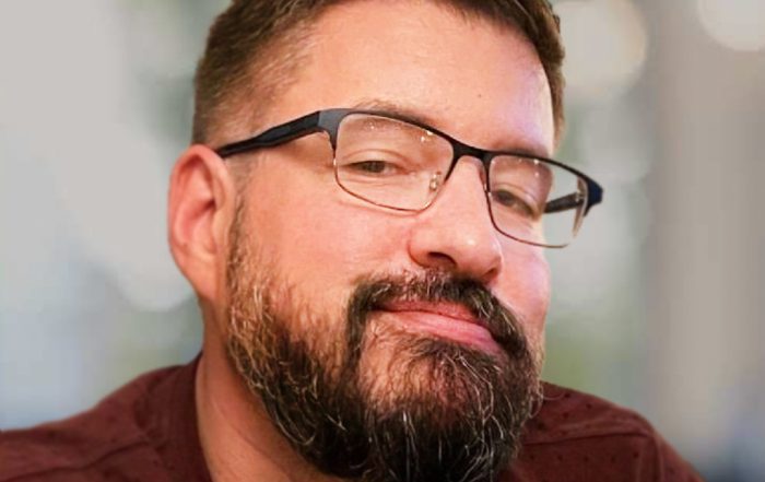 Brando wearing a maroon shirt with a subtle dotted pattern and black-framed glasses, seated indoors. The background is softly blurred, showing neutral tones and diffused light, suggesting a professional or casual setting.