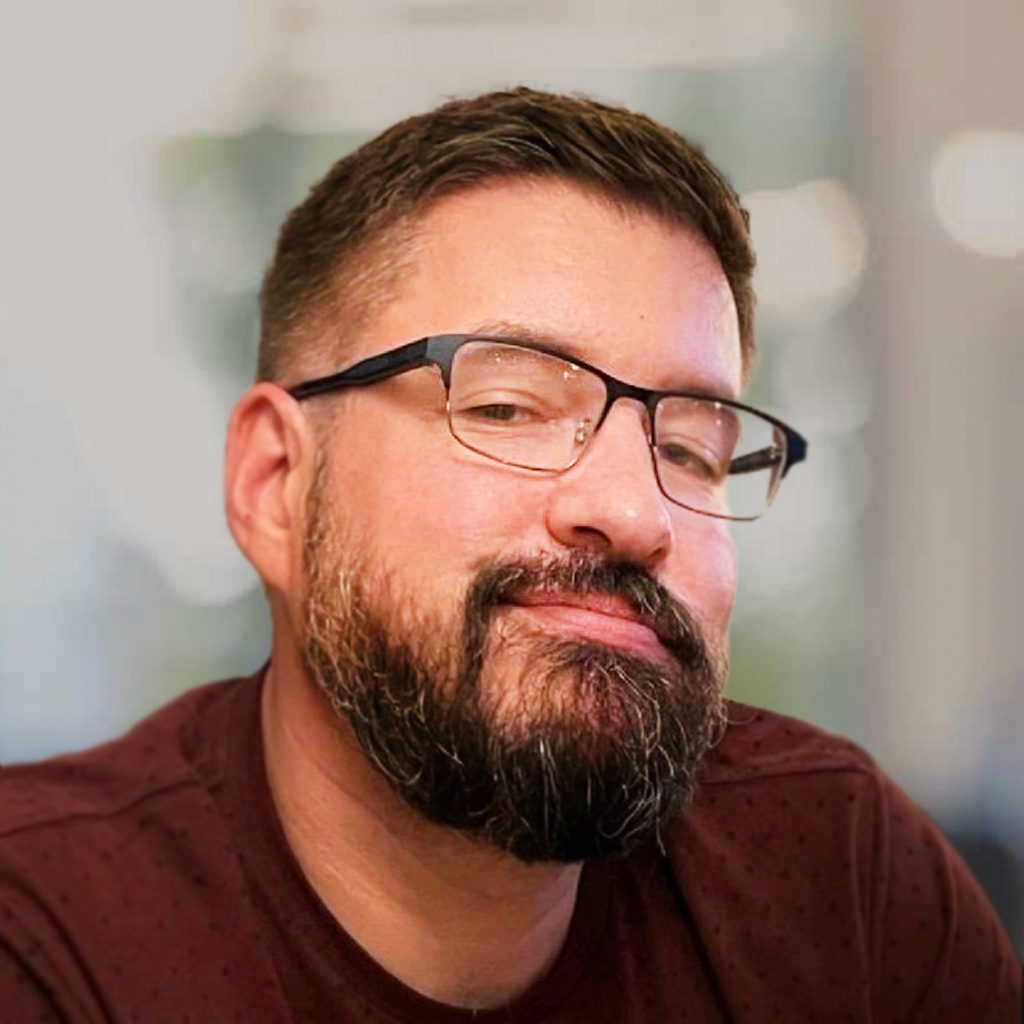 Brando wearing a maroon shirt with a subtle dotted pattern and black-framed glasses, seated indoors. The background is softly blurred, showing neutral tones and diffused light, suggesting a professional or casual setting.