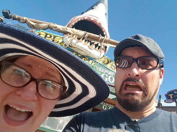 Brando H. and his wife pose playfully in front of a large shark sculpture with its mouth open, mounted above a sign. Brando’s wife wears a wide-brimmed striped hat, while Brando wears a T-shirt and baseball hat. The background shows clear blue sky.