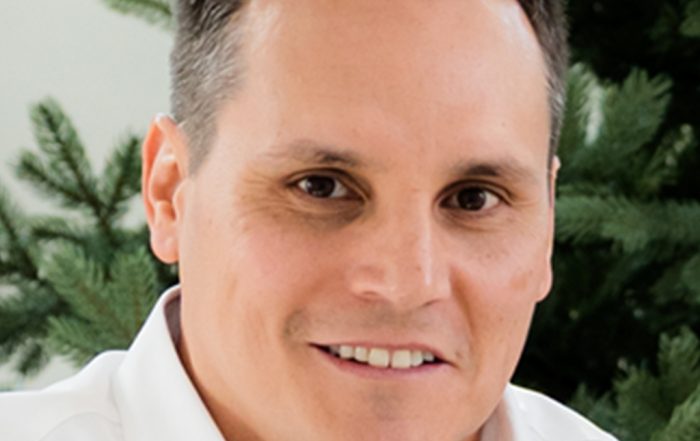 Greg L. wearing a white collared shirt is seated in front of a backdrop of green pine tree branches.