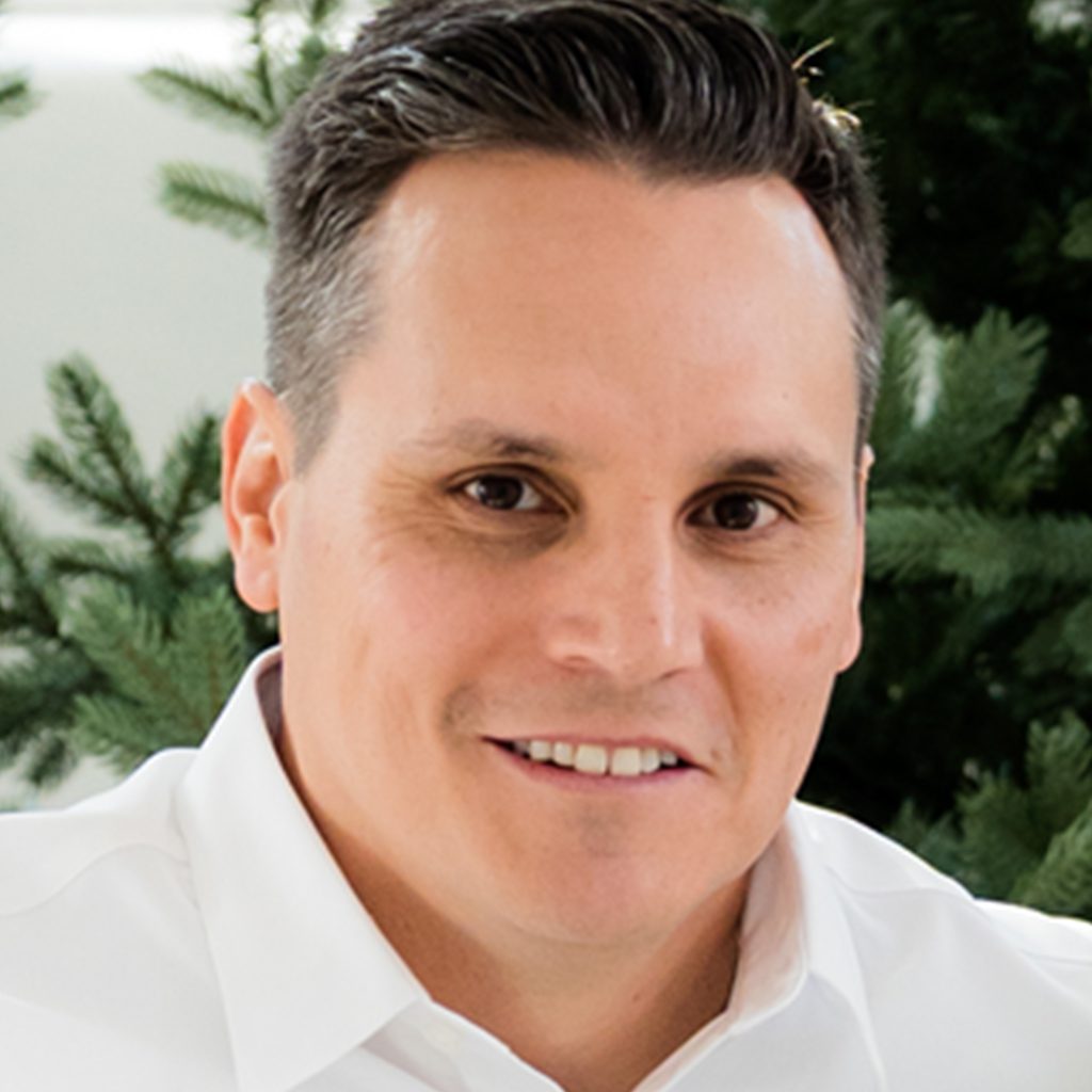Greg L. wearing a white collared shirt is seated in front of a backdrop of green pine tree branches.