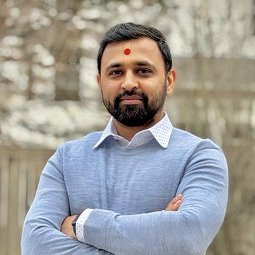 Neel P. standing outdoors with arms crossed, wearing a light blue sweater over a collared shirt, with a wooden fence and trees in the background.