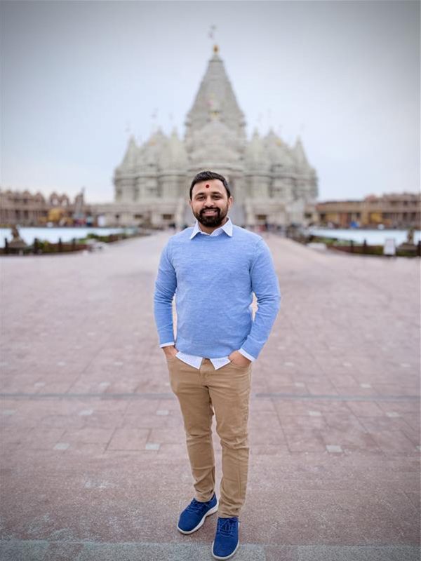 Neel P. standing on a wide walkway in front of the BAPS Swaminarayan Akshardham in Robbinsville, NJ wearing a light blue sweater, khaki pants, and blue shoes.