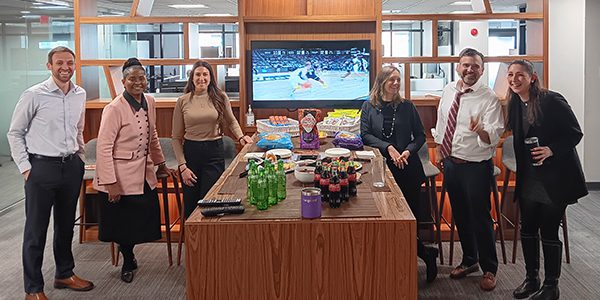 Eight DC crew stand around a tall wooden table in an office common area with snacks and drinks arranged on the tabletop. Shelving, glass walled rooms, and a television screen are visible in the background.
