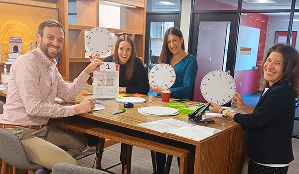 Five DC crew members stand and sit around a tall wooden table in an office workspace, holding paper clocks while assembling volunteer kits. Office shelves, chairs, and glass walled rooms are visible in the background.