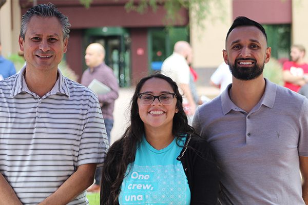 Three people standing side by side outdoors at a campus community event, smiling, with other attendees visible in the background.
