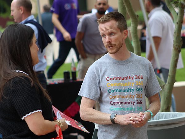 Two people talking at an outdoor Community 365 table during the Arizona Fall Festival with other attendees and trees in the background.