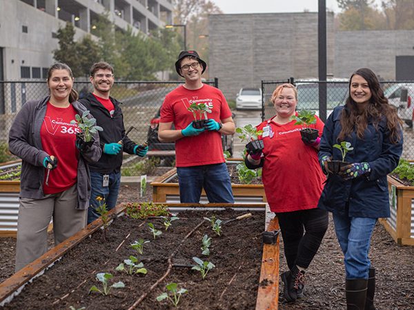 Five crew members stand in raised garden beds holding small plants. The group wears gloves and casual outdoor clothing, with garden plots, fencing, and nearby buildings visible in the background.