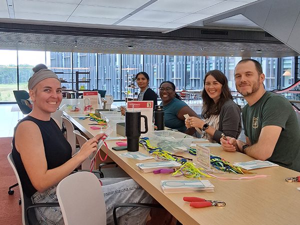Five crew members sit at a long table indoors, working on a hands on volunteer project with craft materials spread across the table. Large windows and a modern office space can be seen in the background.