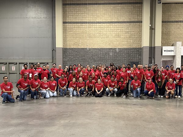 A large group of crew members wearing matching red shirts pose together inside a warehouse style space. The group stands in rows against a tall concrete wall, with signage and doors visible in the background.