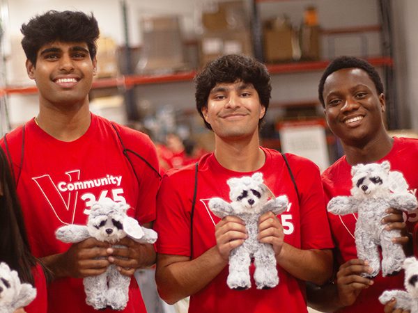 Five Vanguard interns wearing red shirts that read “Community 365” stand in a warehouse space, the Launch Zone, each holding a small stuffed animal. Storage shelves and boxes are visible in the background.
