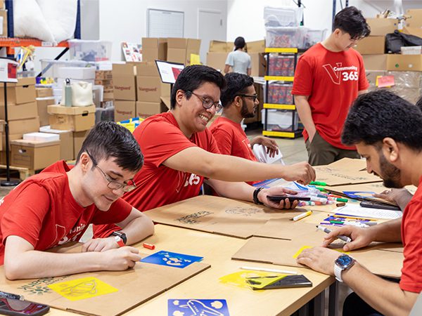 Five Vanguard interns wearing red shirts that read “Community 365” stand in a warehouse space, the Launch Zone, each holding a small stuffed animal. Storage shelves and boxes are visible in the background.
