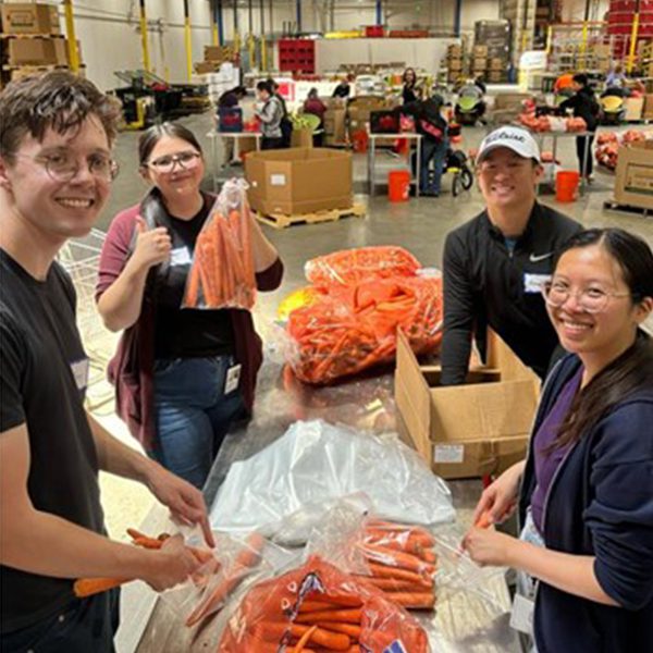 Five volunteers stand around a packing table in a large warehouse, sorting and bagging carrots.