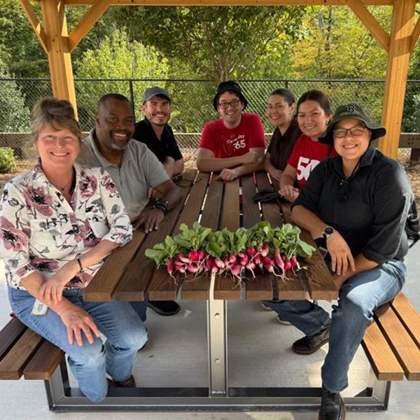 Seven crew members sit around a wooden picnic table under a covered outdoor structure. A small bundle of freshly harvested radishes is displayed on the table, with trees and a fence visible in the background.