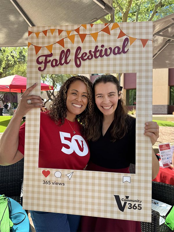 Two crew holding a Fall Festival photo frame with ‘Community 365’ branding while standing outdoors at a campus event