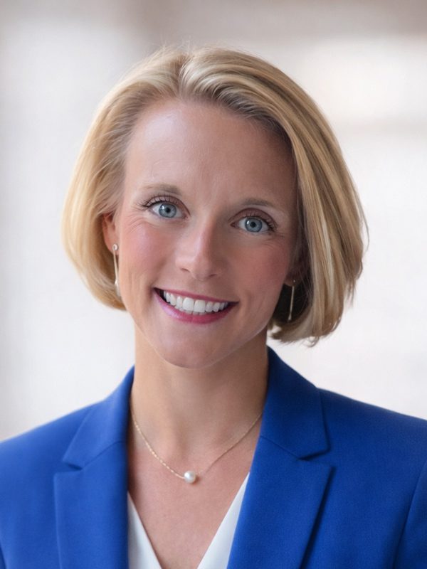 Emily E. smiles confidently wearing a bright blue blazer over a white blouse, posed in front of a softly lit, neutral background.