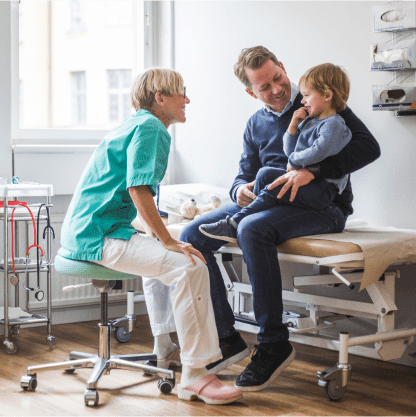 Father holding his young son in a doctor's exam room while a smiling doctor talks to the child.