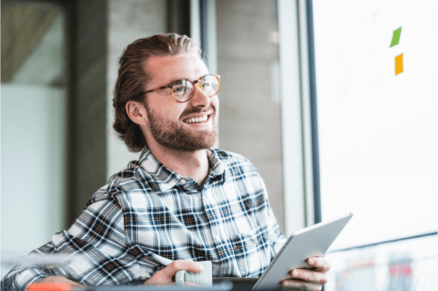 Man looking away from the camera while smiling, looking content, and holding a tablet and coffee mug.