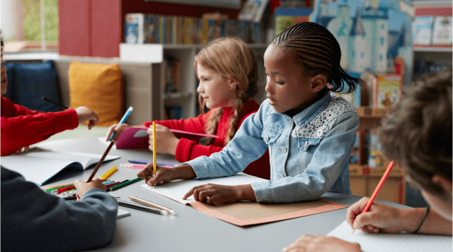 A diverse group of young children sitting at a table in a classroom, coloring and writing.
