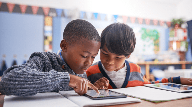 Two young students sitting at a table in a classroom, looking at a tablet