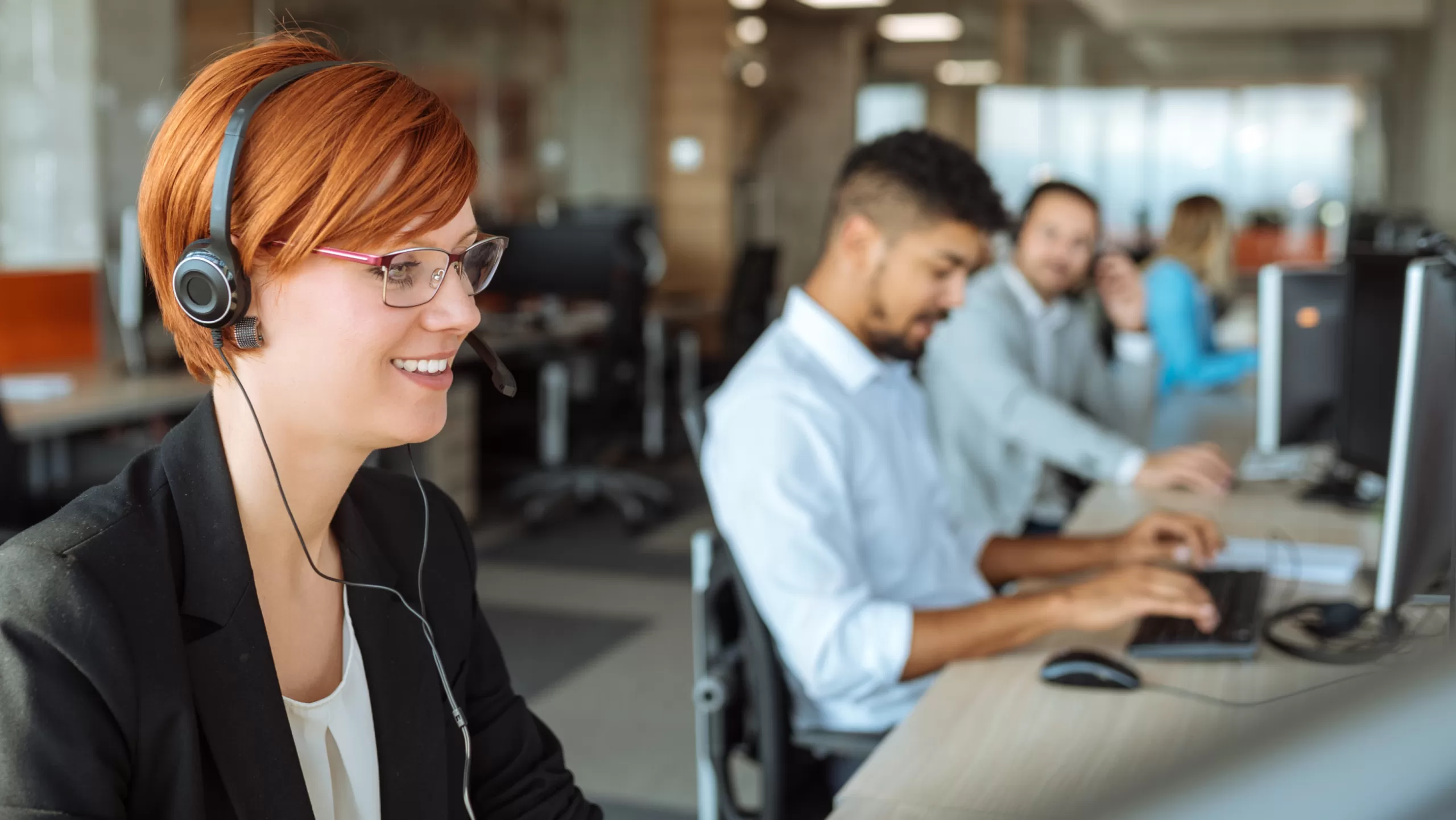 A woman wearing a headset while working on a computer. Several coworkers are out of focus in the background. The coworkers are also working on computers.