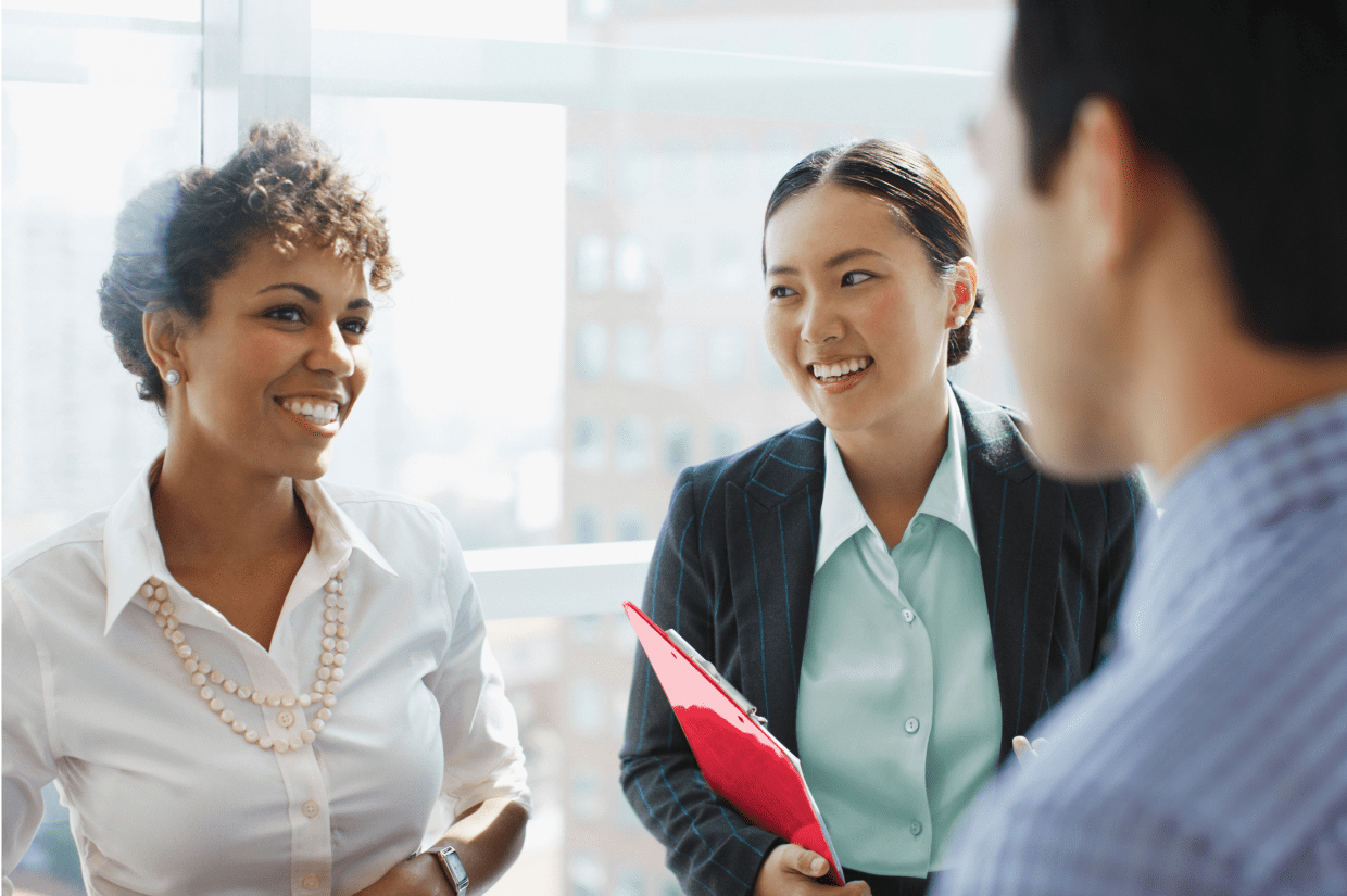 Three coworkers standing and conversing in front of an office window. The woman in the middle holds a red clipboard.