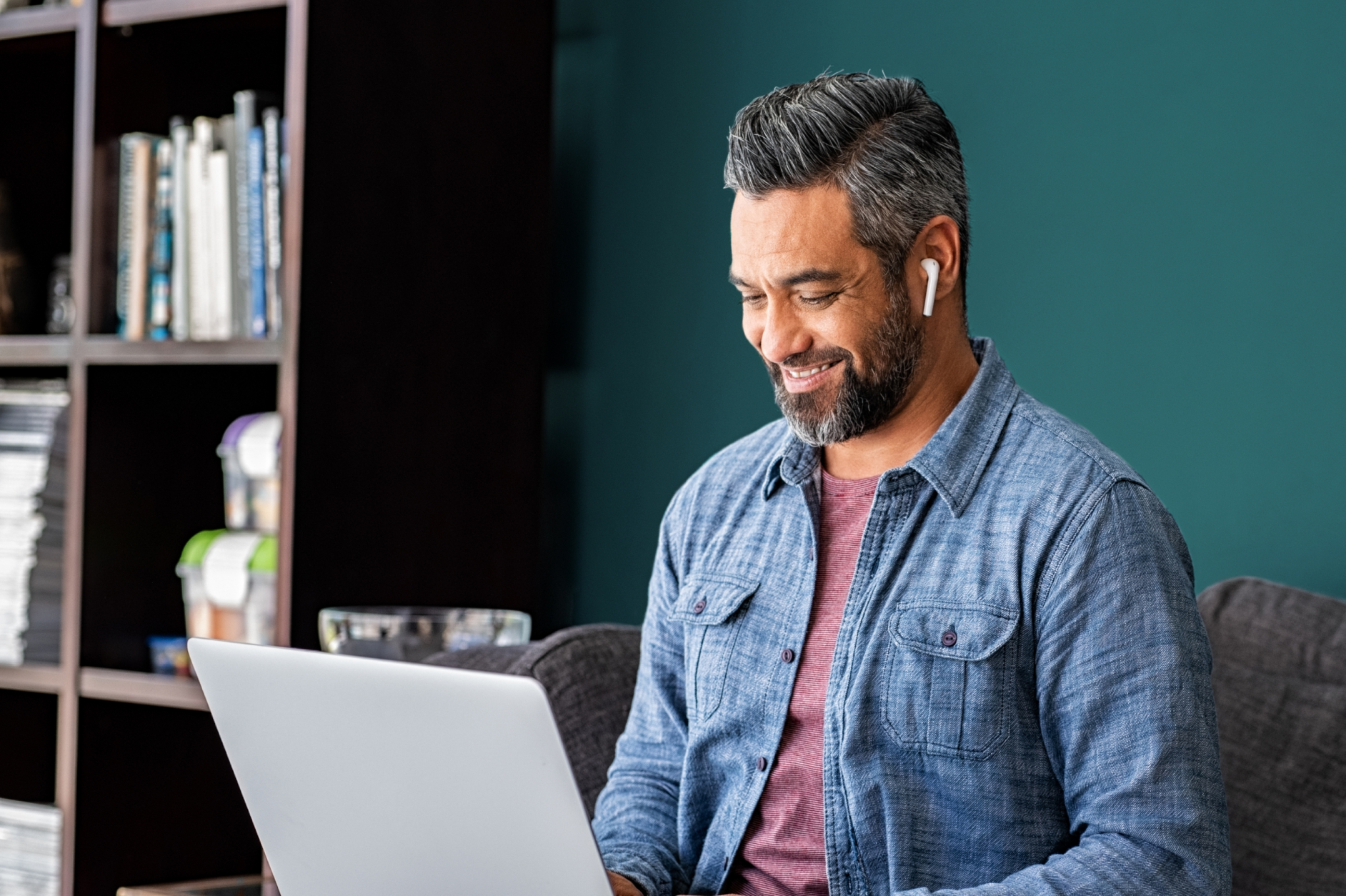 A man working from home. He is sitting on a couch with his open laptop on his lap and he is wearing earbuds.