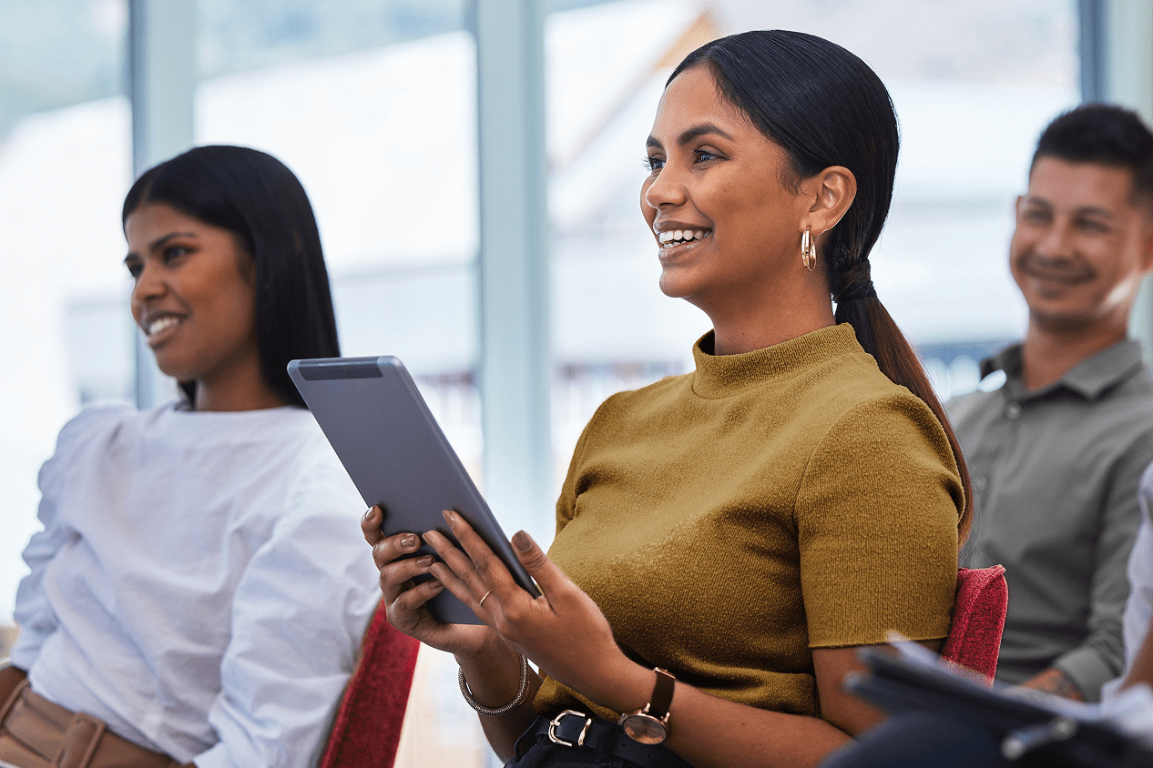 A person seated in the audience at a business presentation, smiling and looking to the right while holding a tablet. Two other audience members are partially visible on the left and right.