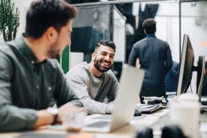 Two young men having a conversation while working on their laptops in an office.