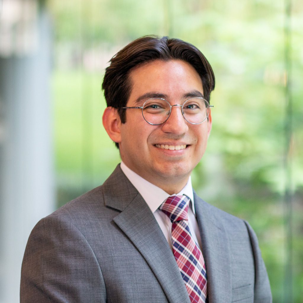 Jose wearing a gray suit jacket, white dress shirt, and a red-and-navy checkered tie, standing indoors with a blurred green outdoor background visible through large glass panels.