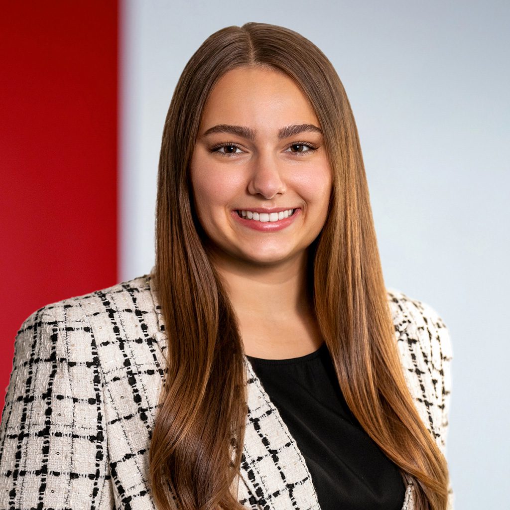 Arianna P. wearing a white and black plaid textured blazer over a black top, posed against a background with a red vertical panel on the left and a light gray section on the right.