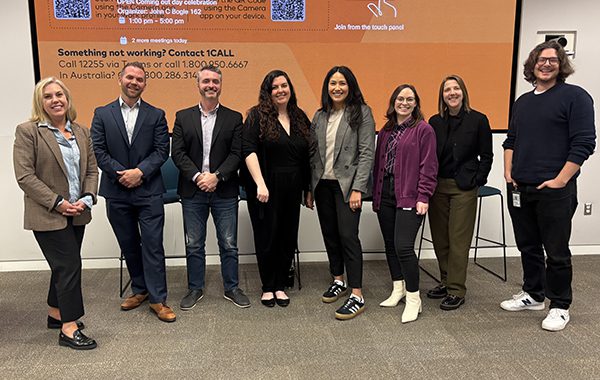 Group of employees standing together in a conference room in front of a large presentation screen with a QR code and event information displayed.