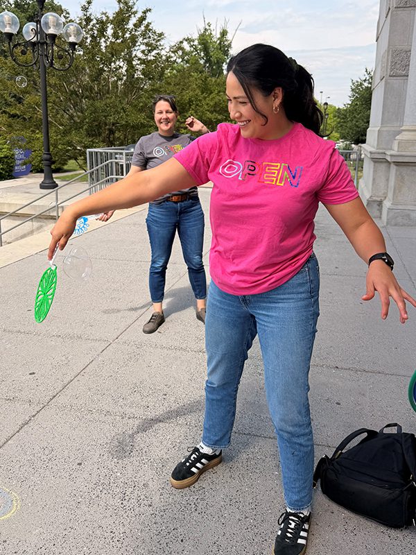 Stephanie A. in a pink OPEN T-shirt playing with a bubble wand outside a building, while another employee stands nearby; chalk drawings and bubble supplies are visible on the ground.