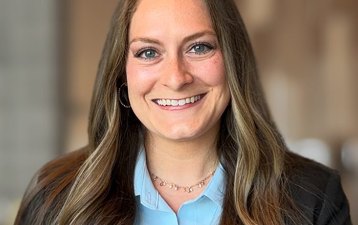 Professional headshot of a person with long brown hair wearing a light blue collared shirt, a dark blazer, hoop earrings, and a delicate necklace. The person is centered in the frame against a softly blurred indoor background